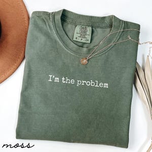 Może przedstawiać: Koszulka w kolorze mchu z białym napisem "I'm the problem". Koszulka ma okrągły dekolt i metkę Comfort Colors. Złoty naszyjnik z wisiorkiem jest zarzucony na koszulkę. Brązowy kapelusz jest widoczny w lewym górnym rogu.