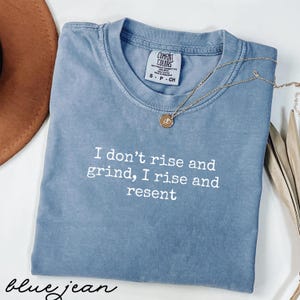 Könnte beinhalten: Blaues Jeans-T-Shirt mit dem weißen Text "I don't rise and grind, I rise and resent". Eine goldene Halskette mit Anhänger liegt über dem Shirt. Das Shirt hat ein Comfort Colors-Etikett.