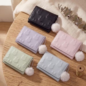 May include: A collection of five small, tri-fold wallets in various pastel colors: black, lavender, pink, mint green, and light blue. Each wallet features a repeating embossed pattern and a fluffy white pom-pom charm attached to a chain.