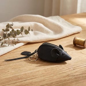 May include: A black leather mouse-shaped coin purse with a zip closure, key ring, and a tail-shaped pull tab. The purse is on a wooden surface with a white cloth and sprig of greenery in the background.