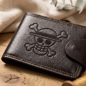May include: A dark brown leather wallet featuring an embossed skull and crossbones emblem. The bi-fold wallet has a snap closure and visible stitching. The wallet is displayed on a wooden surface. The design is reminiscent of a pirate flag.