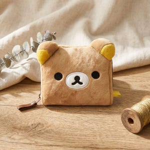 May include: A brown plush wallet shaped like a bear's face, with black eyes and a white and black nose. The bear wallet has small, rounded ears with yellow accents. A brown zipper pull and a spool of gold thread are also visible.