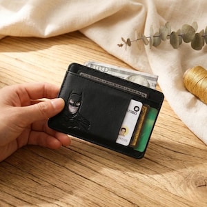 Puede incluir: Tarjetero de cuero negro con diseño de Batman. La cartera tiene múltiples ranuras para tarjetas y un bolsillo para efectivo. El logotipo de Batman está grabado en la parte delantera. El tarjetero se muestra con tarjetas y efectivo.