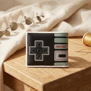 May include: Black wallet with a white trim, styled after a retro games controller. The design includes a directional pad and the word "SELECT" in red. The colour scheme features black, white, and mint green.