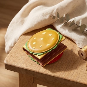 May include: A novelty wallet in the shape of a hamburger. It has a yellow bun with white seed details, green lettuce, a brown patty, and a red base. The wallet is made of leather and rests on a wooden surface.