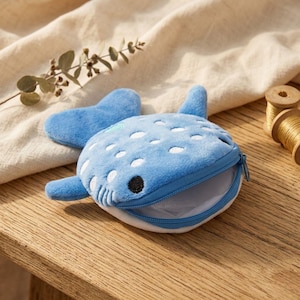 Könnte beinhalten: Eine blaue, walförmige Geldbörse mit weißen Punkten und einem schwarzen Auge. Die Geldbörse hat einen Reißverschluss und ist geöffnet, wodurch das Innere sichtbar wird. Der Hintergrund umfasst eine Holzoberfläche und einen beigen Stoff.