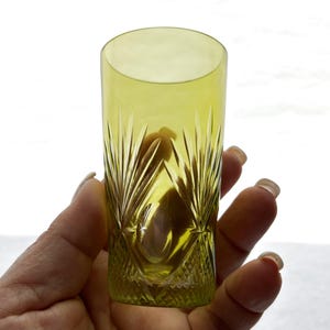 May include: A tall, yellow-green glass with a cut crystal design. The glass has a cylindrical shape and is held in a hand. The cut crystal pattern features vertical lines and diamond shapes, creating a decorative effect. The glass is likely used for drinking.