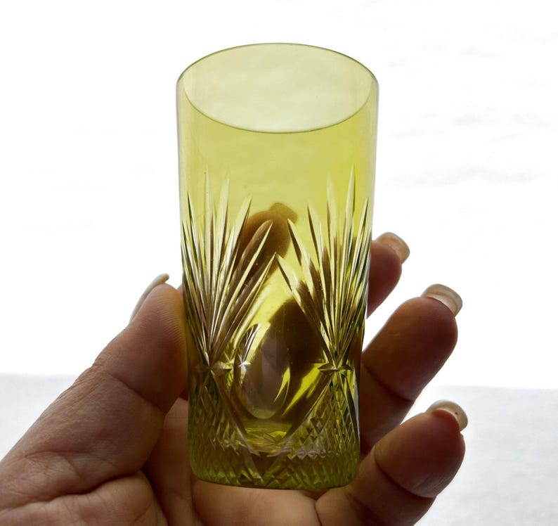 May include: A tall, yellow-green glass with a cut crystal design. The glass has a cylindrical shape and is held in a hand. The cut crystal pattern features vertical lines and diamond shapes, creating a decorative effect. The glass is likely used for drinking.