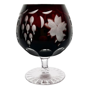 Può includere: Un bicchiere da brandy in cristallo bordeaux con disegni incisi di fiori e grappoli d'uva. Il bicchiere ha un'ampia coppa, un gambo corto e una base decorativa. I disegni incisi sono bianchi.