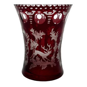 May include: A ruby red glass vase with a detailed etched design. The vase features a deer and forest scene, with a scalloped rim and circular cutouts near the top. The vase is a decorative item.