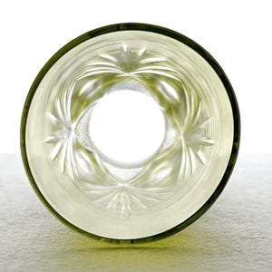 May include: A clear, etched glass with a decorative floral pattern. The glass has a light green tint and a dark green rim. The design features a central circle surrounded by radiating leaf-like shapes. The glass is set against a white background.