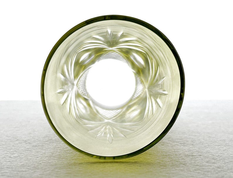 May include: A clear, etched glass with a decorative floral pattern. The glass has a light green tint and a dark green rim. The design features a central circle surrounded by radiating leaf-like shapes. The glass is set against a white background.