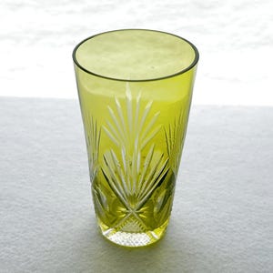 May include: A tall, olive-green glass with a cut-glass design. The glass features a pattern of radiating lines and diamond shapes, creating a textured appearance. The glass is empty and sits on a white surface.