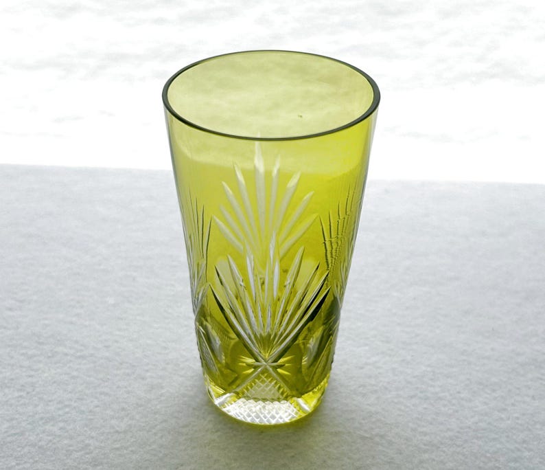 May include: A tall, olive-green glass with a cut-glass design. The glass features a pattern of radiating lines and diamond shapes, creating a textured appearance. The glass is empty and sits on a white surface.