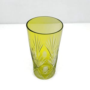 May include: A tall, yellow-green glass with a cut-glass design. The glass has a slightly flared shape and intricate patterns near the base, with vertical lines and leaf-like details. The glass is empty and sits on a white surface.