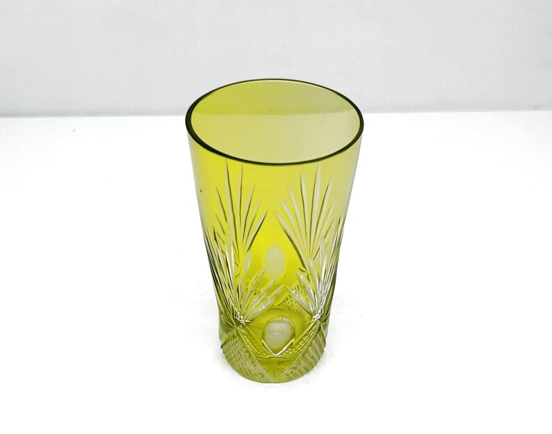 May include: A tall, yellow-green glass with a cut-glass design. The glass has a slightly flared shape and intricate patterns near the base, with vertical lines and leaf-like details. The glass is empty and sits on a white surface.