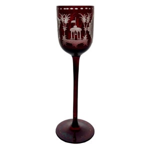 Puede incluir: Un vaso alto y transparente con un tono rojo intenso. El vaso está grabado con un diseño blanco de una pagoda y palmeras. El vaso tiene un tallo largo y una base ancha.