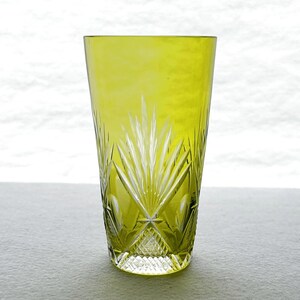 May include: A tall, chartreuse-colored glass with intricate cut-glass designs. The glass features a geometric pattern at the base and a radiating fan-like design extending upwards. The glass is likely used for beverages.