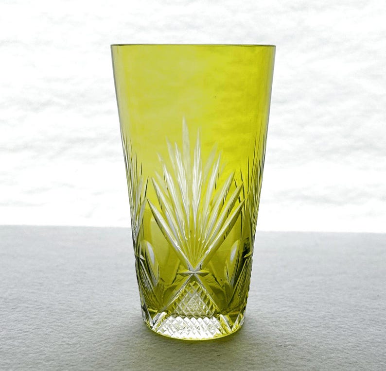 May include: A tall, chartreuse-colored glass with intricate cut-glass designs. The glass features a geometric pattern at the base and a radiating fan-like design extending upwards. The glass is likely used for beverages.