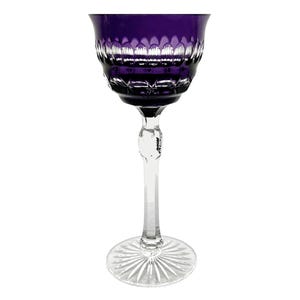 May include: A deep purple glass goblet featuring a wide bowl with a textured, scalloped pattern near the rim. The stem is clear and faceted, leading down to a circular base that displays a radial, sunburst-like cut pattern etched into the glass.