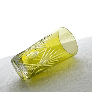 May include: A yellow-green, cut-glass tumbler with a geometric and fan-like pattern. The glass is cylindrical with a slightly flared rim. The tumbler is lying on a white surface, showcasing its intricate design and color.