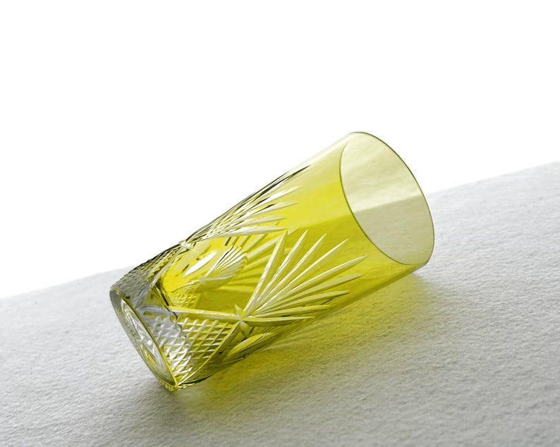 May include: A yellow-green, cut-glass tumbler with a geometric and fan-like pattern. The glass is cylindrical with a slightly flared rim. The tumbler is lying on a white surface, showcasing its intricate design and color.