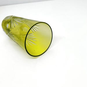 May include: A yellow-green glass tumbler with etched starburst and leaf designs. The glass has a thick, black rim and is lying on its side. The glass is likely used for drinking beverages.