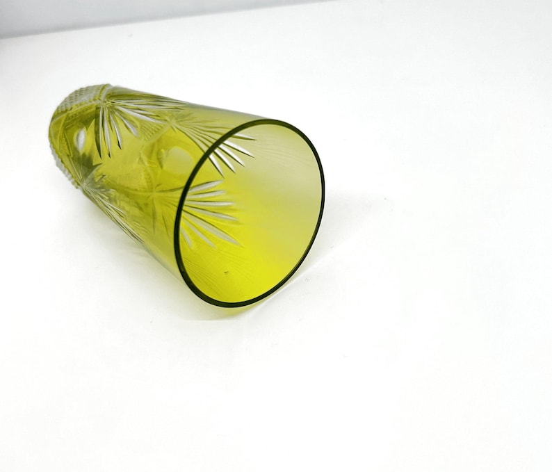 May include: A yellow-green glass tumbler with etched starburst and leaf designs. The glass has a thick, black rim and is lying on its side. The glass is likely used for drinking beverages.