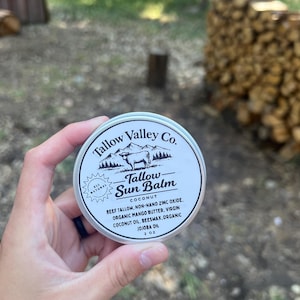 Op de afbeelding: Een ronde zilveren metalen blik van Tallow Valley Co. Tallow Sun Balm met een wit etiket met een zwarte illustratie van een berg en een koe. Het etiket vermeldt ingrediënten zoals rundvet, niet-nano zinkoxide, mangoboter en kokosolie, 57 g.