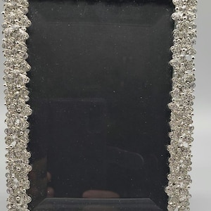 May include: Silver-toned rectangular picture frame with a black interior. The frame is adorned with a floral design, embellished with sparkling rhinestones. A decorative item for displaying photos or artwork.