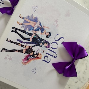 May include: A white rectangular box featuring a printed graphic of three anime-style characters in varied outfits, the name Sofia in dark blue text, and two decorative purple satin bows attached to the corners of the lid.