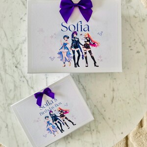 May include: Two white rectangular gift boxes featuring a purple satin bow on top. The larger box displays the name Sofia and an illustration of three anime-style characters. The smaller box is labeled Sofia's Birthday Box with the same characters.