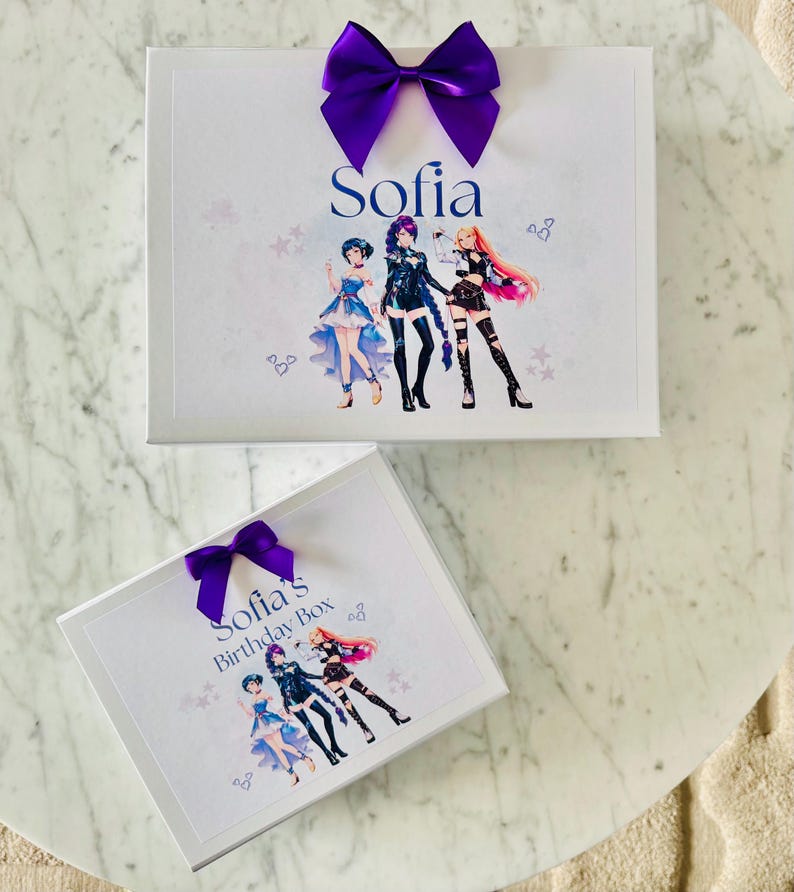 May include: Two white rectangular gift boxes featuring a purple satin bow on top. The larger box displays the name Sofia and an illustration of three anime-style characters. The smaller box is labeled Sofia's Birthday Box with the same characters.