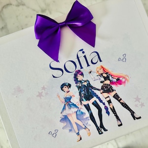 May include: A white rectangular gift box featuring a large purple satin bow at the top. The name Sofia is printed in dark blue script. Below the name are three anime-style characters in colorful outfits, surrounded by small heart and star icons.