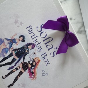 May include: A white rectangular gift box featuring a purple satin ribbon bow. The lid displays the text Sofia's Birthday Box in blue, accompanied by an illustration of three anime-style characters and small gray star and heart graphics.