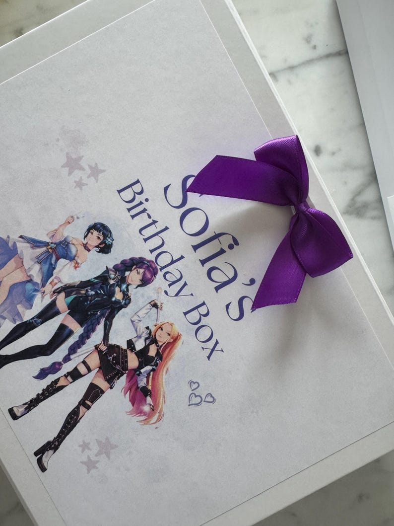 May include: A white rectangular gift box featuring a purple satin ribbon bow. The lid displays the text Sofia's Birthday Box in blue, accompanied by an illustration of three anime-style characters and small gray star and heart graphics.