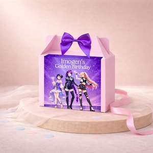 May include: A light pink gable box featuring a purple satin bow on top. The front panel displays the text Imogen's Golden Birthday above an illustration of three anime-style characters in colorful outfits against a sparkling purple background.