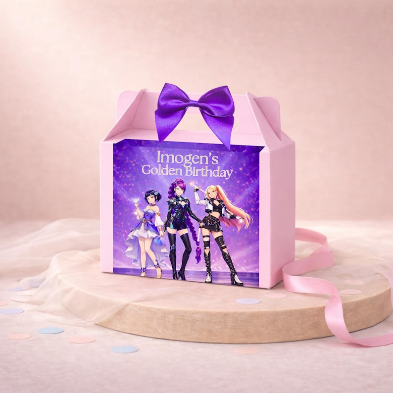 May include: A light pink gable box featuring a purple satin bow on top. The front panel displays the text Imogen's Golden Birthday above an illustration of three anime-style characters in colorful outfits against a sparkling purple background.