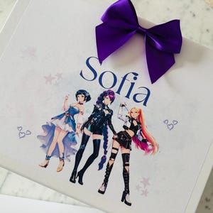 May include: A white rectangular gift box featuring a large purple satin bow at the top. The box is printed with the name Sofia in dark blue script, accompanied by a colorful illustration of three anime-style characters and small heart and star icons.