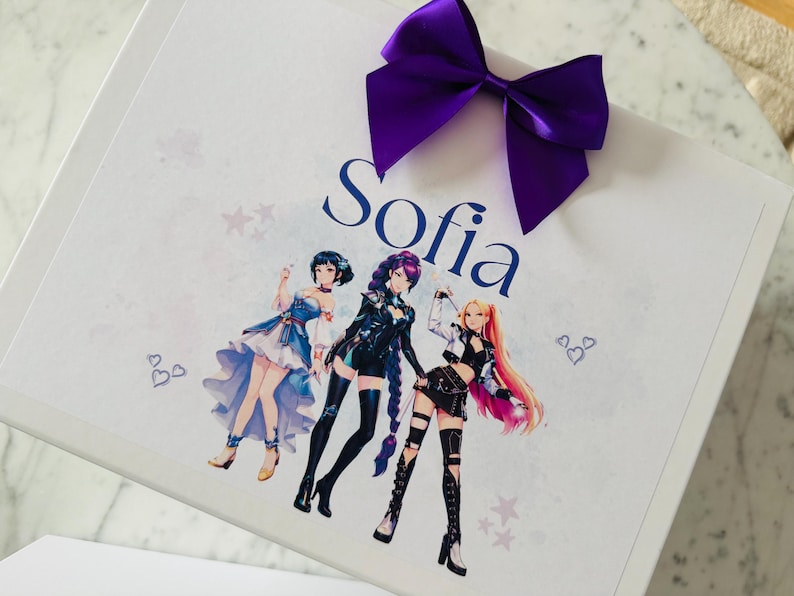 May include: A white rectangular gift box featuring a large purple satin bow at the top. The box is printed with the name Sofia in dark blue script, accompanied by a colorful illustration of three anime-style characters and small heart and star icons.