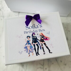 May include: A white rectangular gift box featuring a purple satin ribbon bow on top. The lid displays the text Sofia's Birthday Box in blue lettering above a colorful illustration of three anime-style characters with distinct outfits and hairstyles.