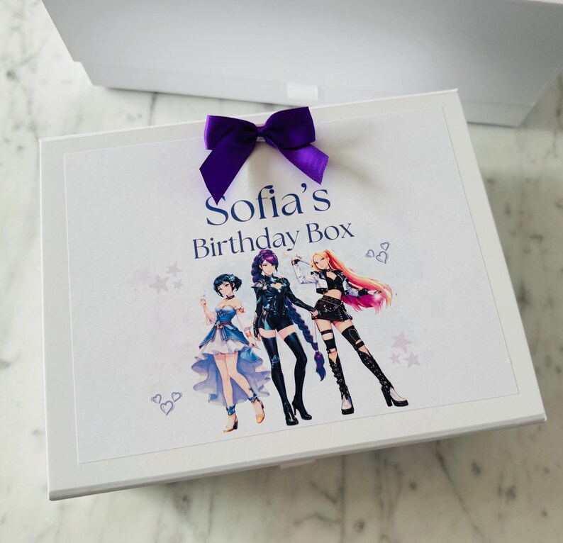 May include: A white rectangular gift box featuring a purple satin ribbon bow on top. The lid displays the text Sofia's Birthday Box in blue lettering above a colorful illustration of three anime-style characters with distinct outfits and hairstyles.