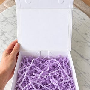 May include: A square white gift box with a hinged lid, featuring a magnetic closure tab at the top. The interior of the box is filled with light purple crinkle-cut paper shred, providing a decorative cushion for contents within the container.