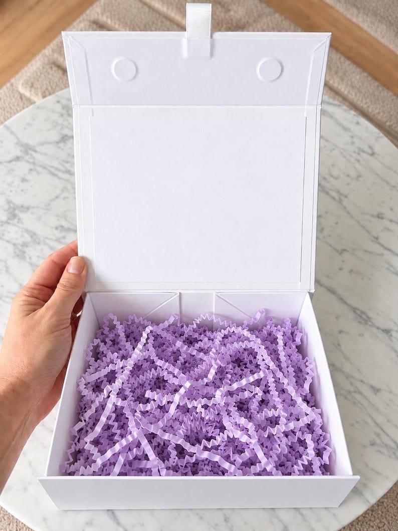 May include: A square white gift box with a hinged lid, featuring a magnetic closure tab at the top. The interior of the box is filled with light purple crinkle-cut paper shred, providing a decorative cushion for contents within the container.