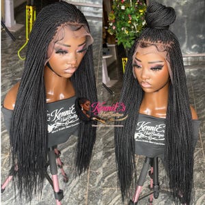 May include: Two black braided wigs on mannequin heads. One wig has long braids, the other has long braids with a bun on top. Both wigs have a lace front and are displayed on a stand. The text "Kennie's Hair Boutique" is visible.