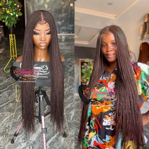 May include: A long, dark brown braided wig on a mannequin head, displayed on a tripod. The wig features a natural-looking hairline and is styled with a center part. The image also shows a person wearing the same wig.