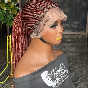 May include: A reddish-brown braided wig styled in a high ponytail. The wig features a lace front and is displayed on a mannequin. The mannequin is wearing a gray off-the-shoulder top with the text "Kenni's Hair Boutique".