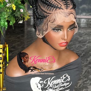May include: A black braided wig styled in a high bun, with intricate cornrow patterns near the hairline. The wig is displayed on a mannequin head, with the "Kenni's Hair Boutique" logo visible on the mannequin's top. The text "Where Braids Meet Convenience" is also visible.