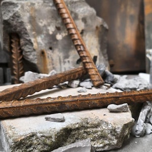 Puede incluir: Primer plano de barras de refuerzo oxidadas sobre bloques de hormigón gris. Las barras de refuerzo tienen una textura estriada y son de color marrón oscuro. La escena sugiere construcción o demolición.