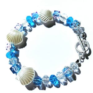 May include: A bracelet featuring three white seashell charms interspersed with faceted blue and clear crystal beads. The jewellery piece is secured with a silver-toned metal toggle clasp, creating a circular arrangement of alternating shell and glass elements.